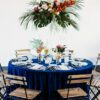 Round table with blue linen. White china and tropical florals. Wood slatted chairs with black metal.