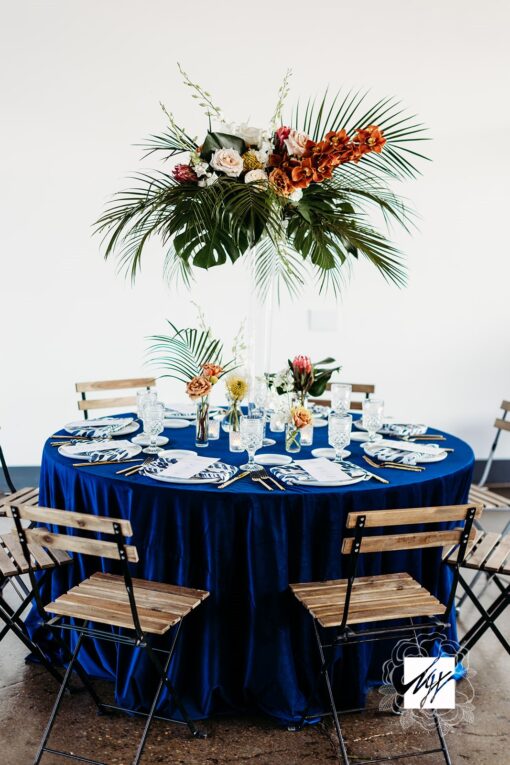 Round table with blue linen. White china and tropical florals. Wood slatted chairs with black metal.