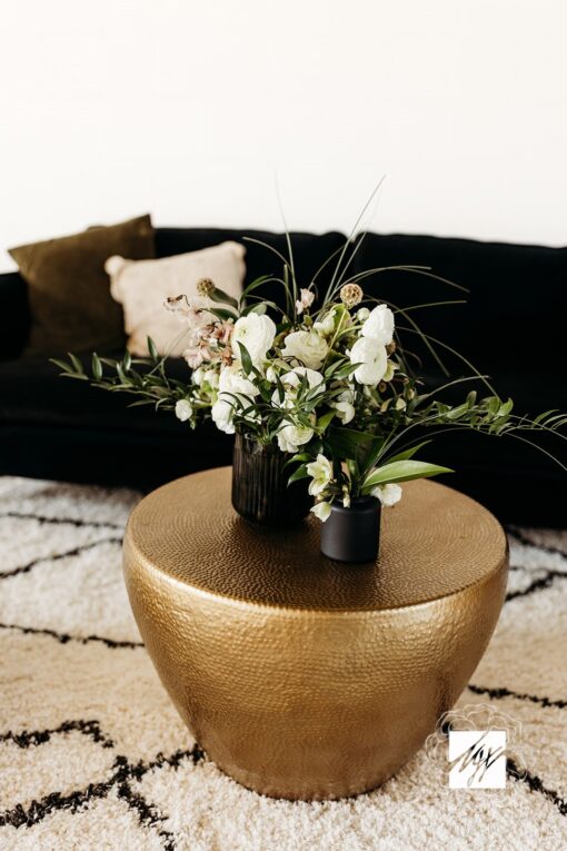 Gold round accent table with florals. Sitting on a black and white rug with a black sofa in the background