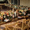 Light wood x-back chairs in a U shape head table with wooden farm tables and lovely florals and tablescape at an outdoor wedding reception dinner.