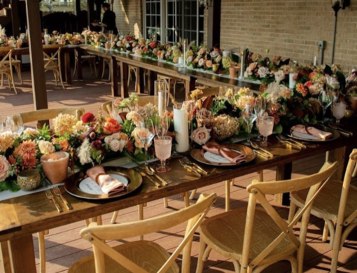 Light wood x-back chairs in a U shape head table with wooden farm tables and lovely florals and tablescape at an outdoor wedding reception dinner.