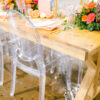 Light wooden farm table with clear acrylic chairs. Bright tablescape with orange, pink, and greens.