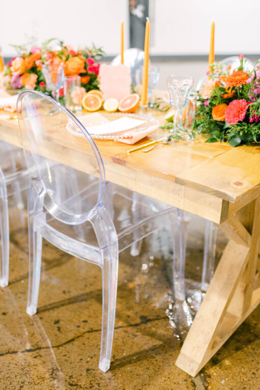 Light wooden farm table with clear acrylic chairs. Bright tablescape with orange, pink, and greens.