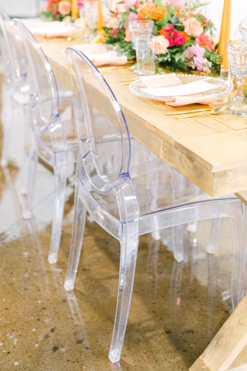 Clear acrylic chairs at a light wood farm table with bright orange and pink tablescape.