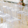 Clear acrylic chairs in a row at an indoor wedding reception. White background. Orange and pink florals