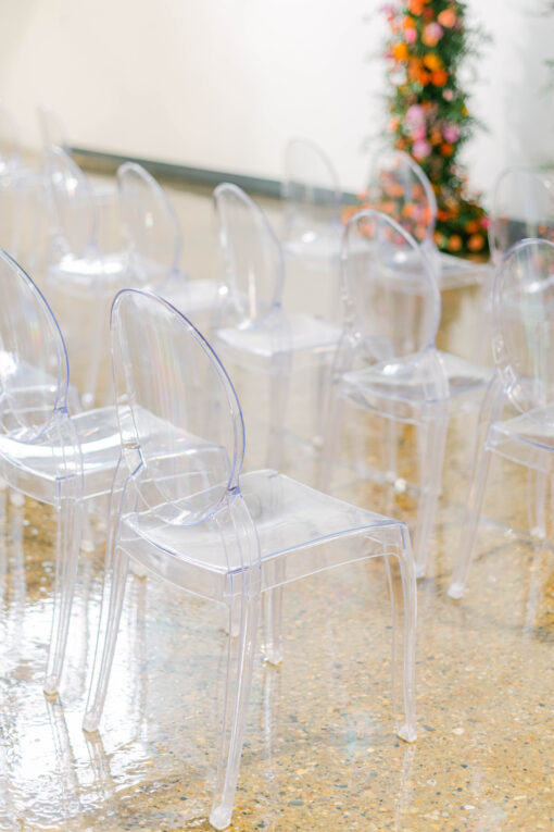 Clear acrylic chairs in a row at an indoor wedding reception. White background. Orange and pink florals