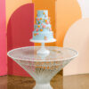 Mid-century modern white fiberglass twisty round table with a light blue wedding cake against orange and pink backdrop