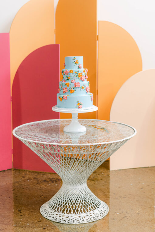 Mid-century modern white fiberglass twisty round table with a light blue wedding cake against orange and pink backdrop