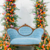 Vintage curved blue velvet sofa set in a backdrop of two floral pillars with bright orange, pink, and green. White background and concrete floor
