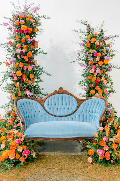 Vintage curved blue velvet sofa set in a backdrop of two floral pillars with bright orange, pink, and green. White background and concrete floor