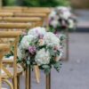 Light colored X-back chairs in rows at an outdoor wedding ceremony. Florals on the ends of each row.