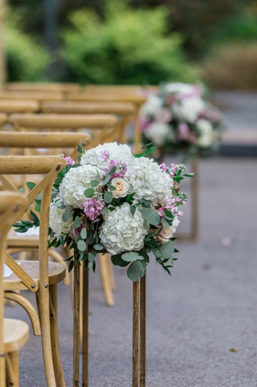 Light colored X-back chairs in rows at an outdoor wedding ceremony. Florals on the ends of each row.