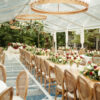 Wedding reception with a clear tent, a long row of farm tables and high end Louis chairs. Circular lighting strung from the ceiling