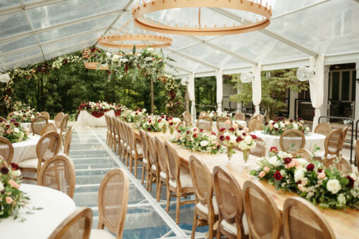 Wedding reception with a clear tent, a long row of farm tables and high end Louis chairs. Circular lighting strung from the ceiling