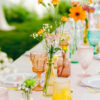 Outdoor wedding reception with peach, yellow, pink, and teal goblets, white plates, and colorful small vases with orange flowers and baby's breath.