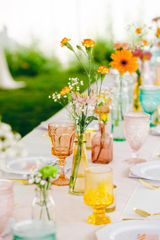 Outdoor wedding reception with peach, yellow, pink, and teal goblets, white plates, and colorful small vases with orange flowers and baby's breath.