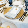 Tablesetting at wedding reception with white linen, gold charger, gold flatware, and navy blue goblets