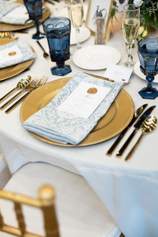 Tablesetting at wedding reception with white linen, gold charger, gold flatware, and navy blue goblets
