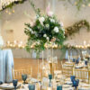 View of a round table at a wedding reception with tall florals and place settings with gold and blue accents.