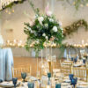 View of a round table at a wedding reception with tall florals and place settings with gold and blue accents.