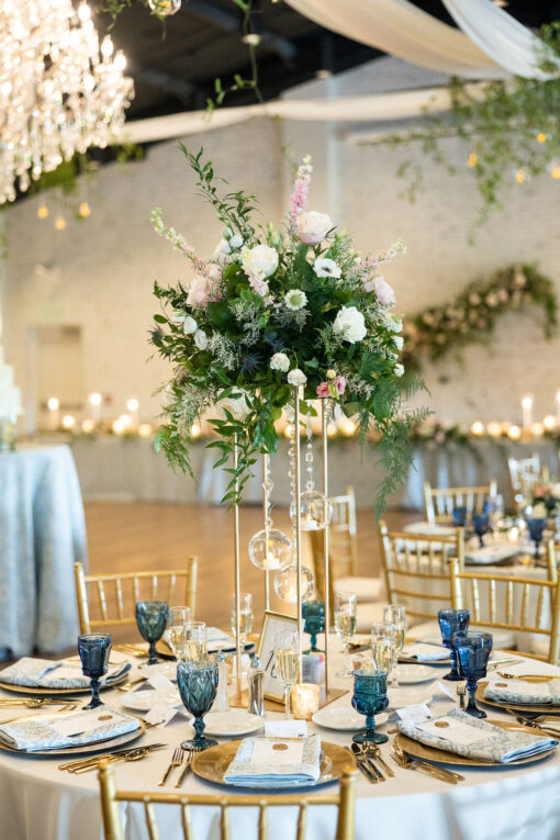 View of a round table at a wedding reception with tall florals and place settings with gold and blue accents.