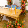 Wooden farm table with a vintage blue settee, two place settings, and candles. Orange and pink florals/