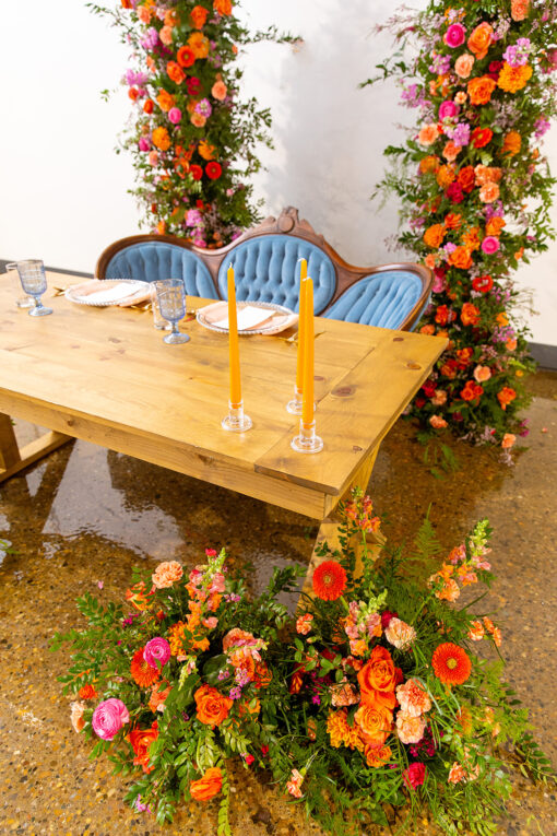Wooden farm table with a vintage blue settee, two place settings, and candles. Orange and pink florals/