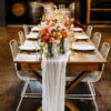 Warm wooden farm table with white wire chairs, orange hues on florals.