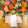 Tablescape with clear plate, light blue goblet, bold pink and orange flowers on a light wood farm table.