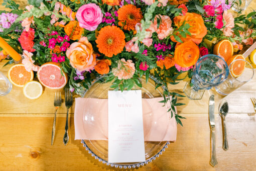 Tablescape with clear plate, light blue goblet, bold pink and orange flowers on a light wood farm table.