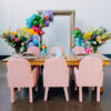 View of the back of soft pink arm chairs against a wooden farm table. Bold colors with florals on the table. Giant frame backdrop with balloons behind the table against a white wall.
