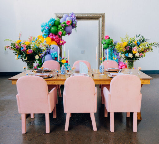 View of the back of soft pink arm chairs against a wooden farm table. Bold colors with florals on the table. Giant frame backdrop with balloons behind the table against a white wall.