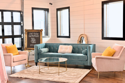 Spring lounge seating area with a modern blue sofa, pink velvet arm chairs on either side with yellow pillows. Cream area rug with a round gold coffee table with glass top. White walls with windows in the background.